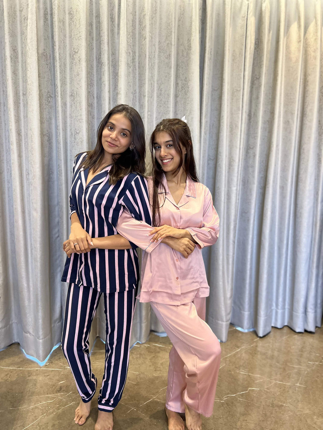 Two women standing barefoot wearing stylish satin pajamas, one in navy and pink stripes and the other in solid light pink, in front of grey curtains