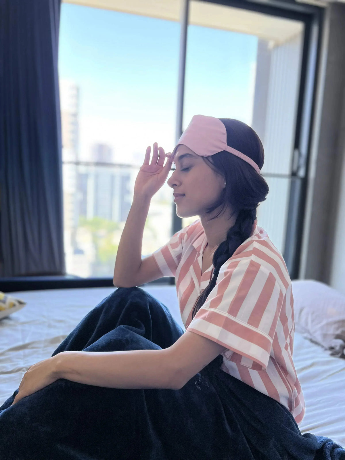 Woman in striped pajamas and pink sleep mask sitting on bed by window in bright bedroom