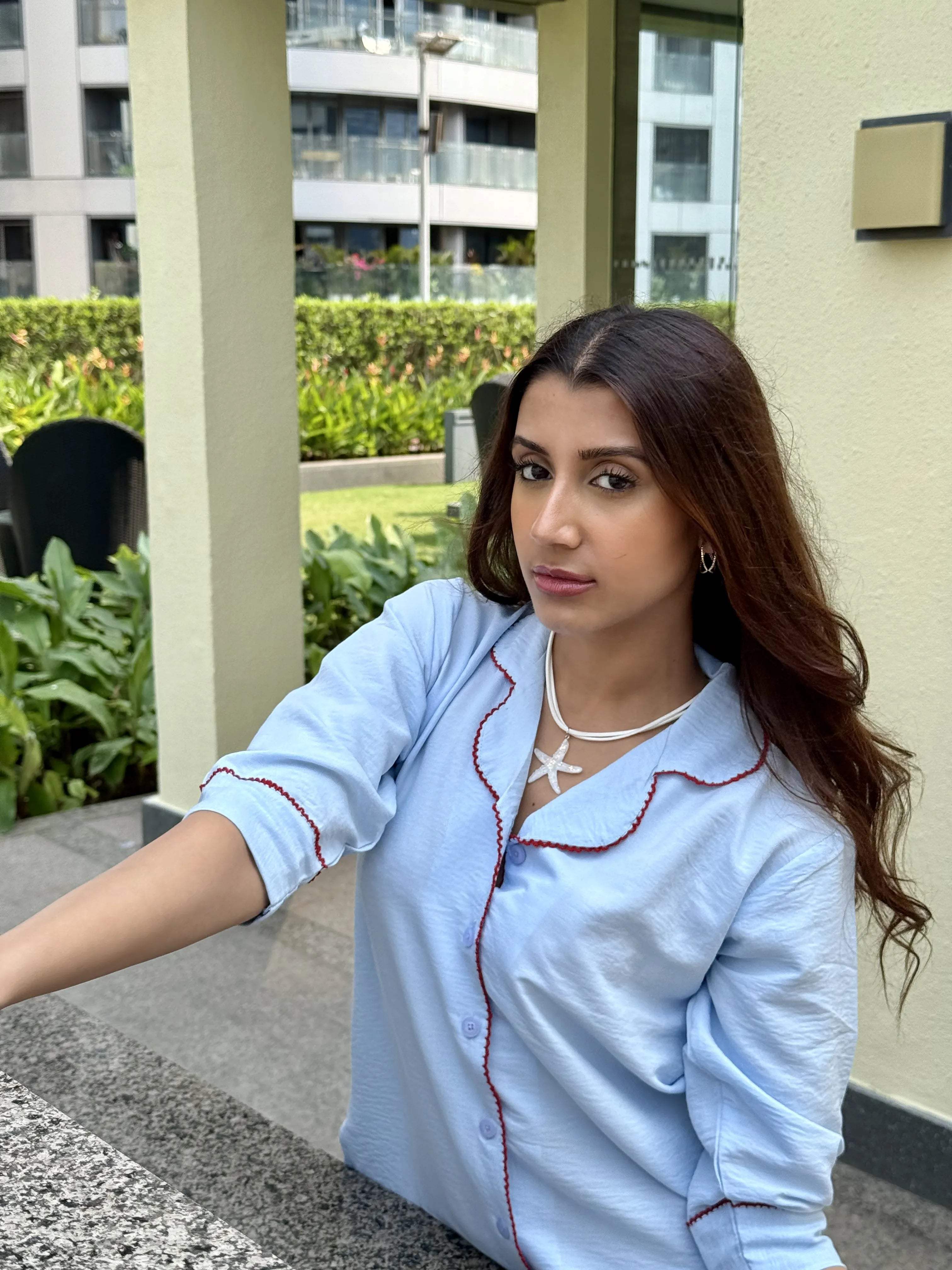 Woman in a light blue pajama top with red trim and starfish necklace outdoors near building and greenery