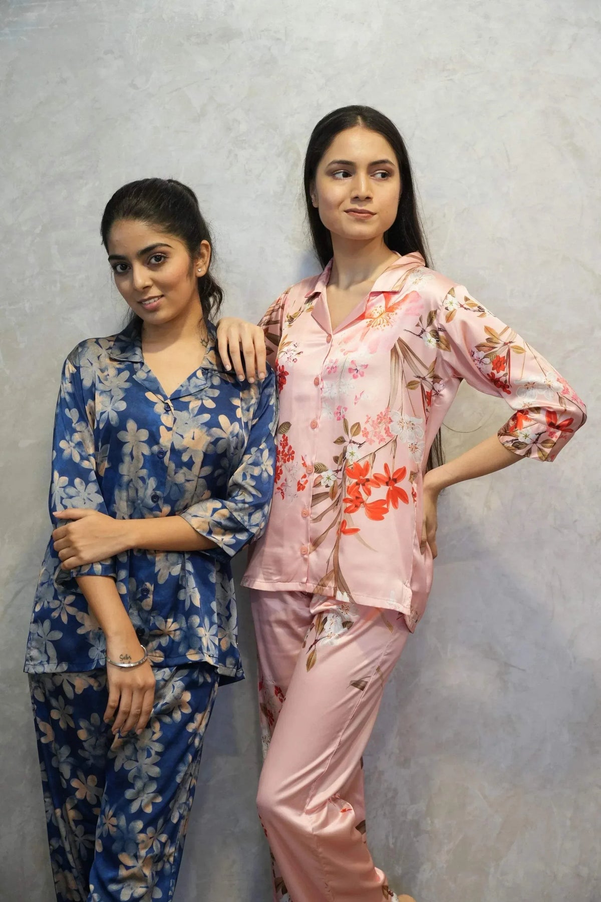 Two women posing in floral printed satin pajamas, one in blue and beige, the other in pink with red flowers