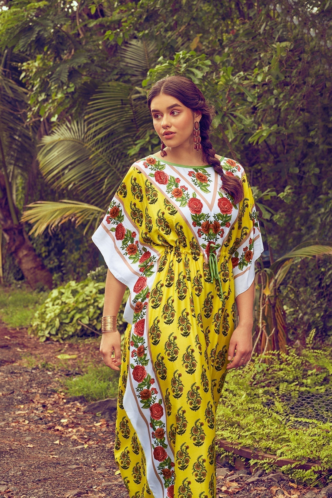 Woman in a vibrant yellow floral maxi dress with pink and green accents standing in a lush garden