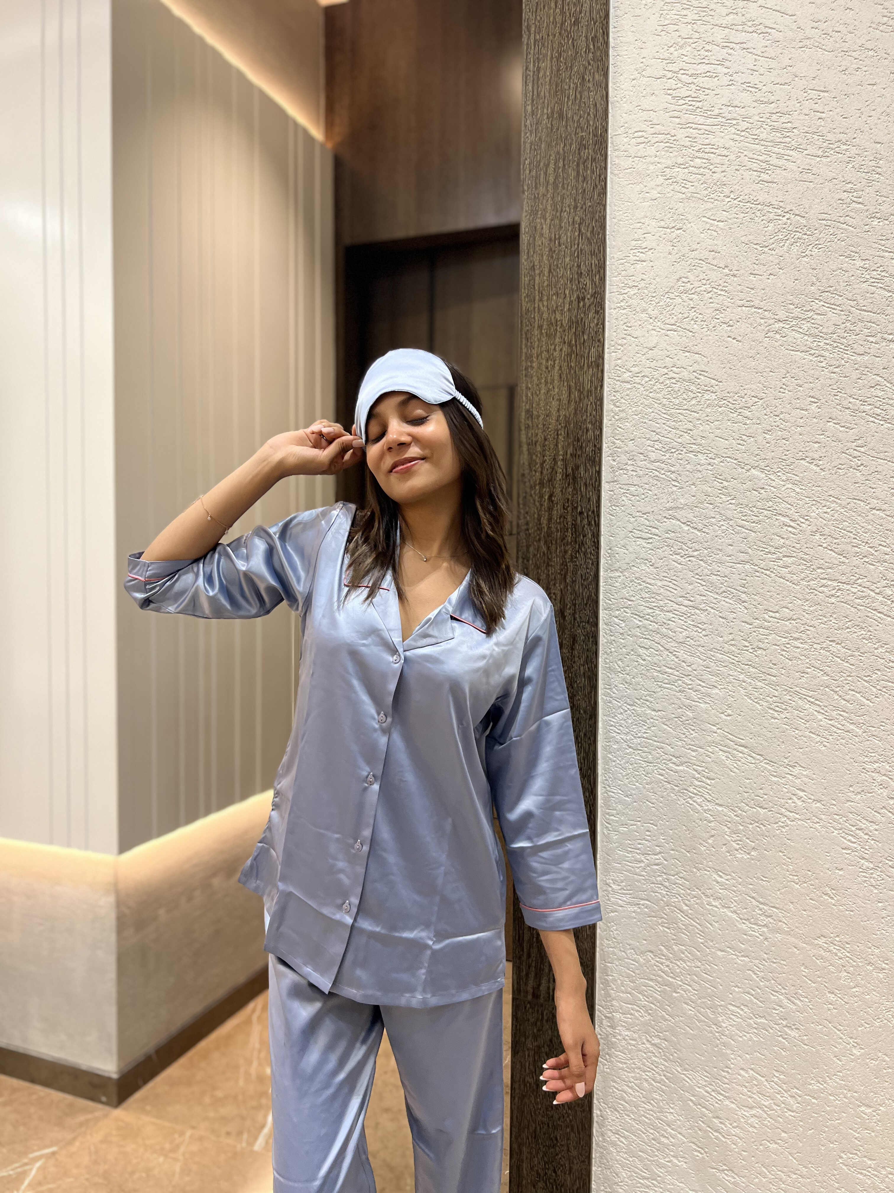 Woman in light blue satin pajamas and matching sleep mask posing indoors against beige and wood wall