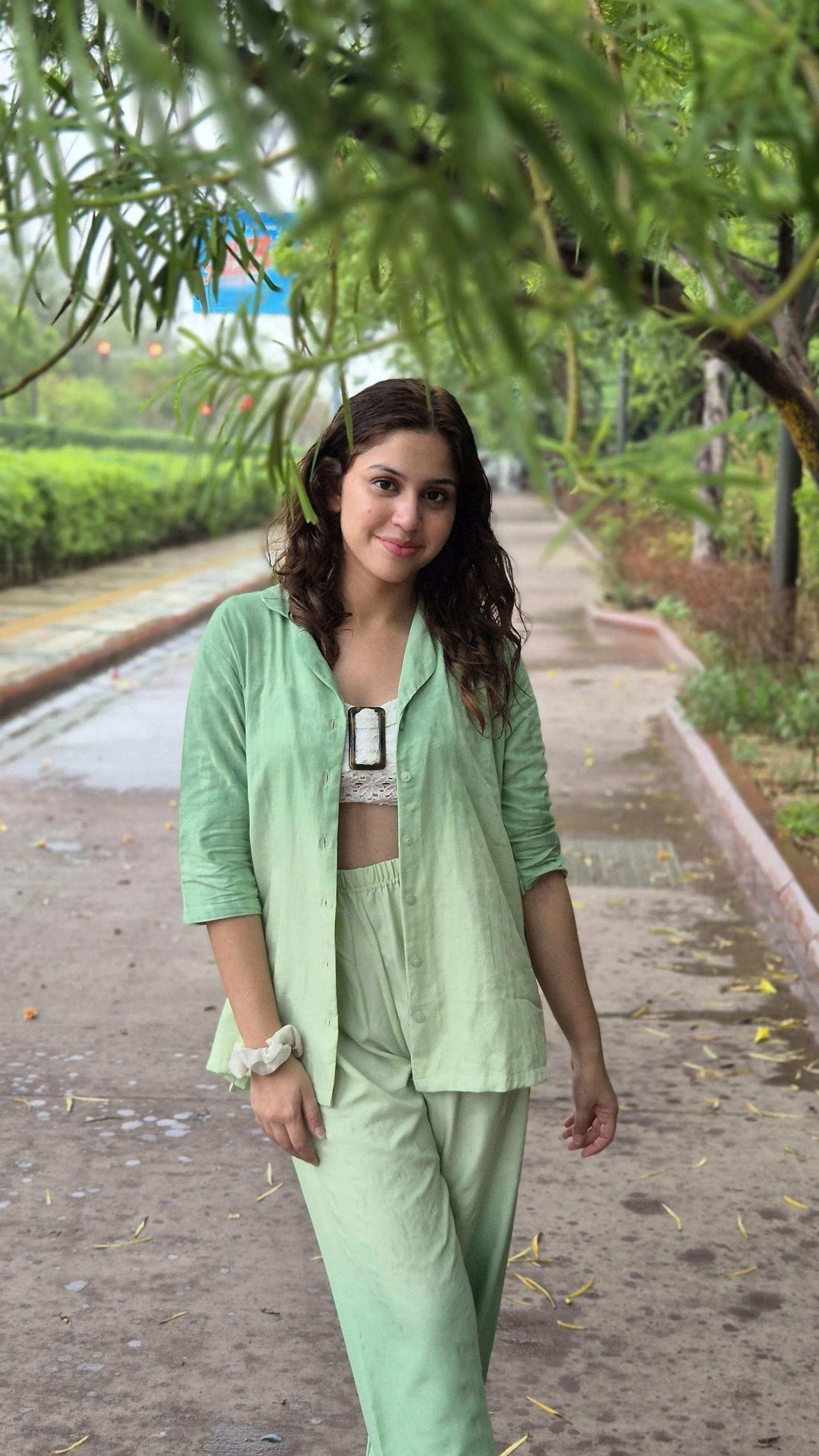 Young woman smiling in casual light green outfit standing on a tree-lined sidewalk on a cloudy day