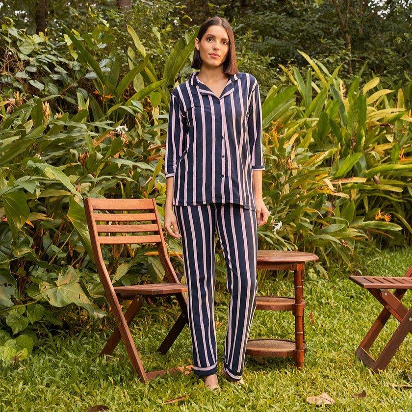 Woman wearing navy blue and pink striped pajamas standing on grass near wooden chairs and table in lush garden