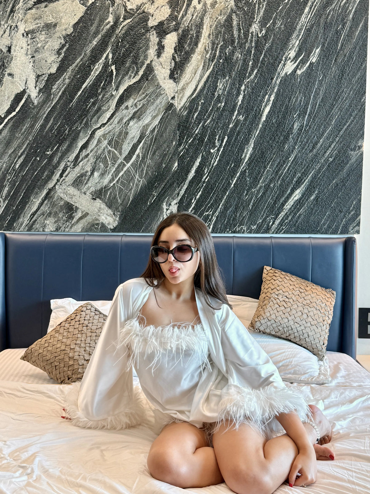 Woman in white feather-trimmed satin robe and sunglasses sitting on bed with blue headboard and decorative pillows