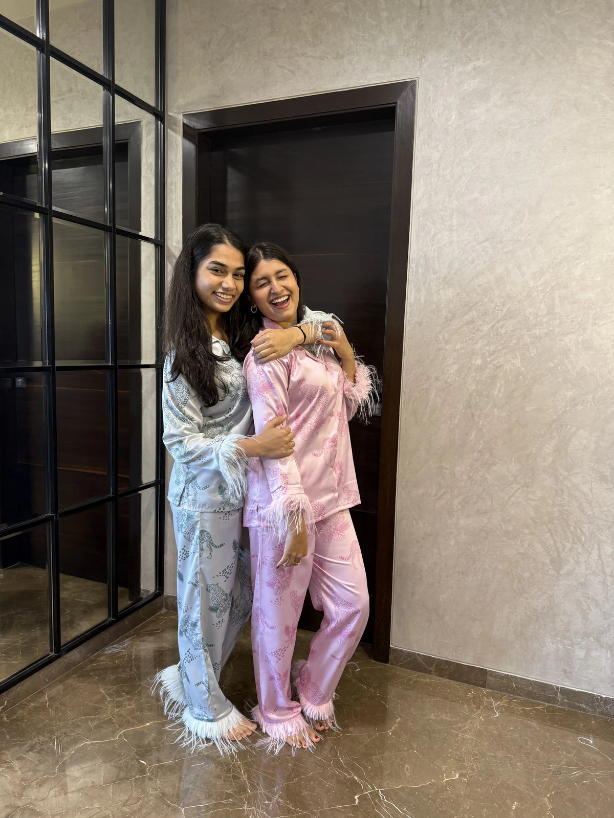 Two women smiling and hugging indoors wearing satin pajamas with feather trim and animal print