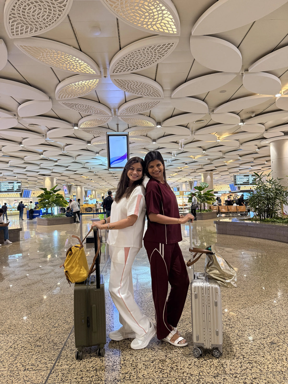 Two women standing back-to-back in an airport terminal with stylish luggage and trendy travel outfits