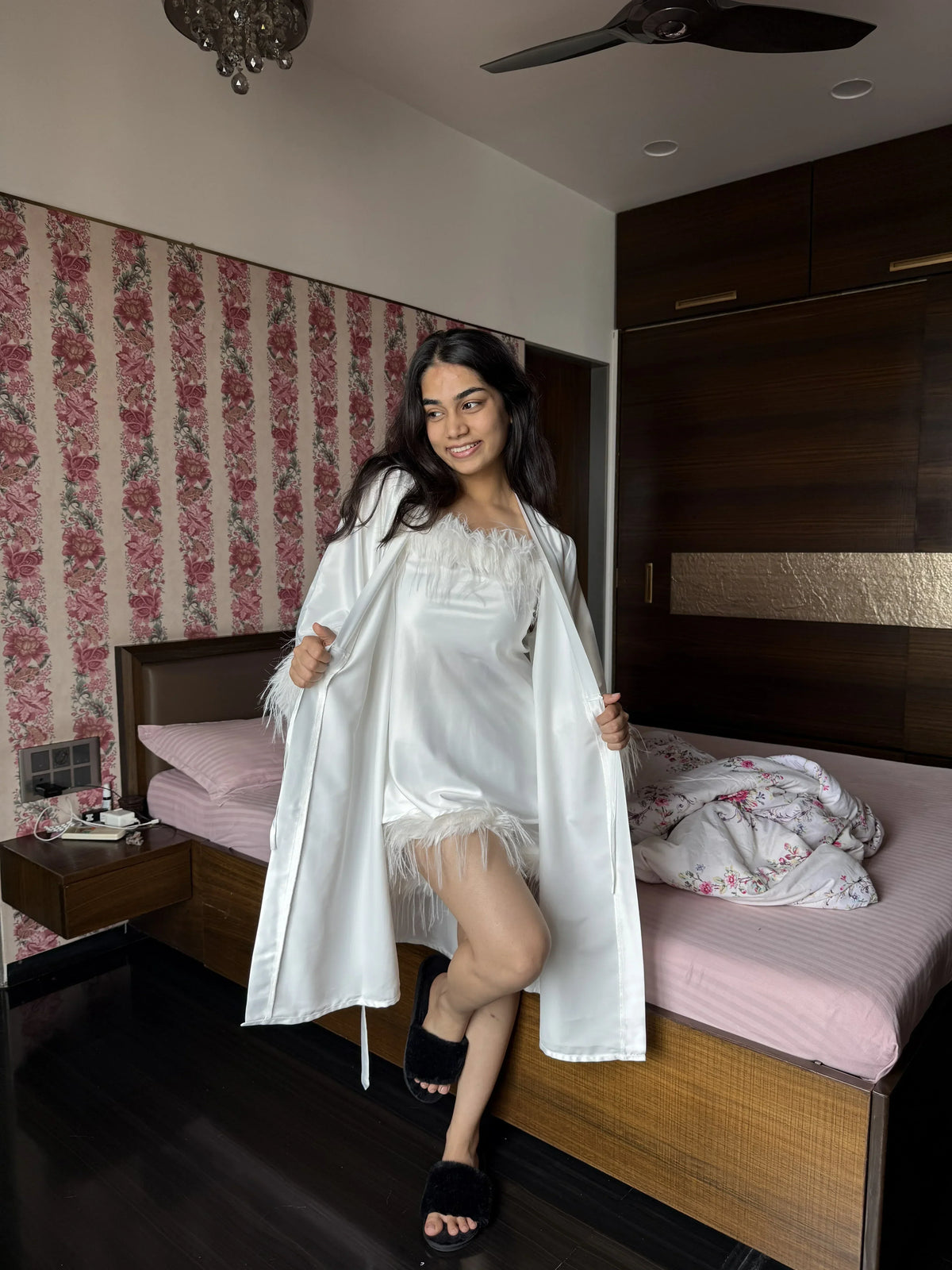 Woman in white satin robe and nightwear posing by bedroom with floral wallpaper and wooden furniture