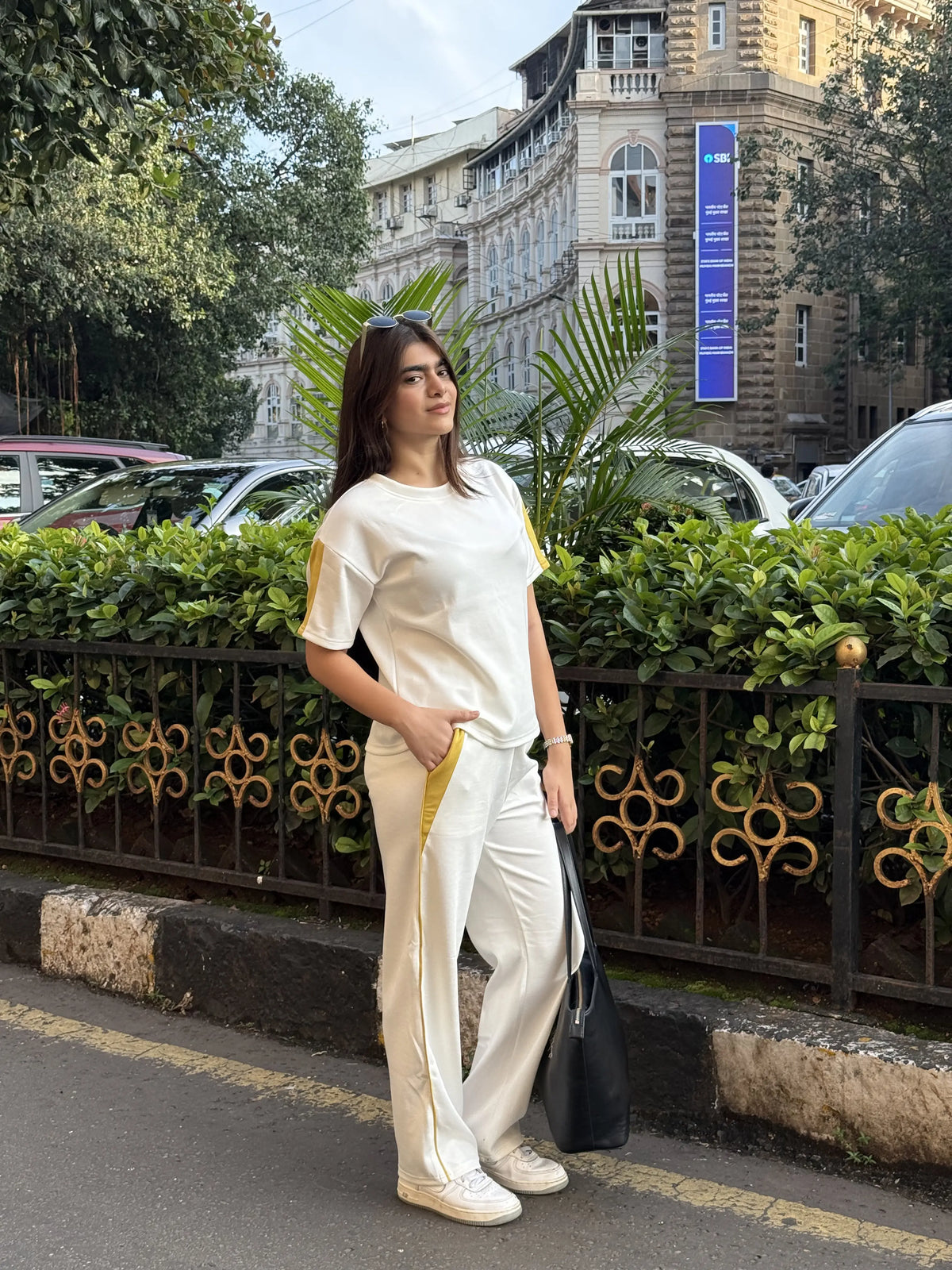 Woman in white casual outfit with yellow accents and sneakers standing on city street with greenery and historic buildings