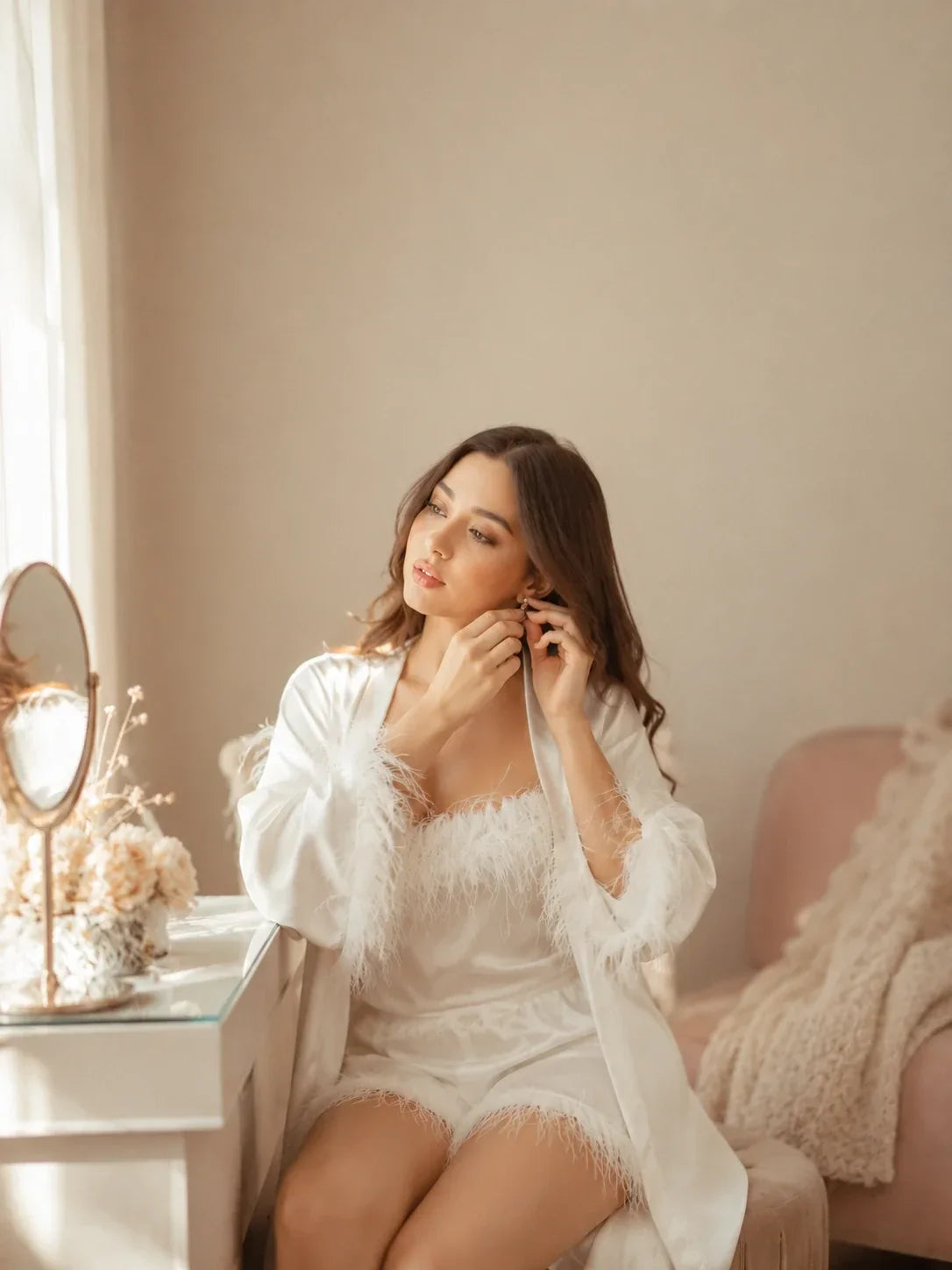 Woman in a white satin feather-trim robe and matching shorts adjusting her earring at a softly lit vanity in a romantic bridal-inspired bedroom.