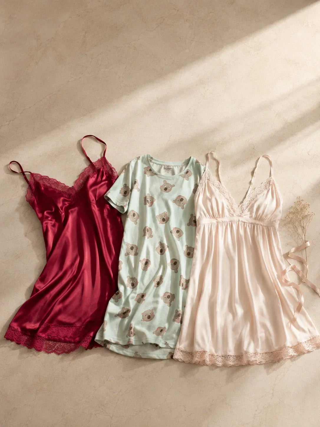 Flat lay of three folded sleepwear styles—a wine satin chemise, printed cotton nightgown, and blush babydoll—on a warm marble surface with dried flowers.