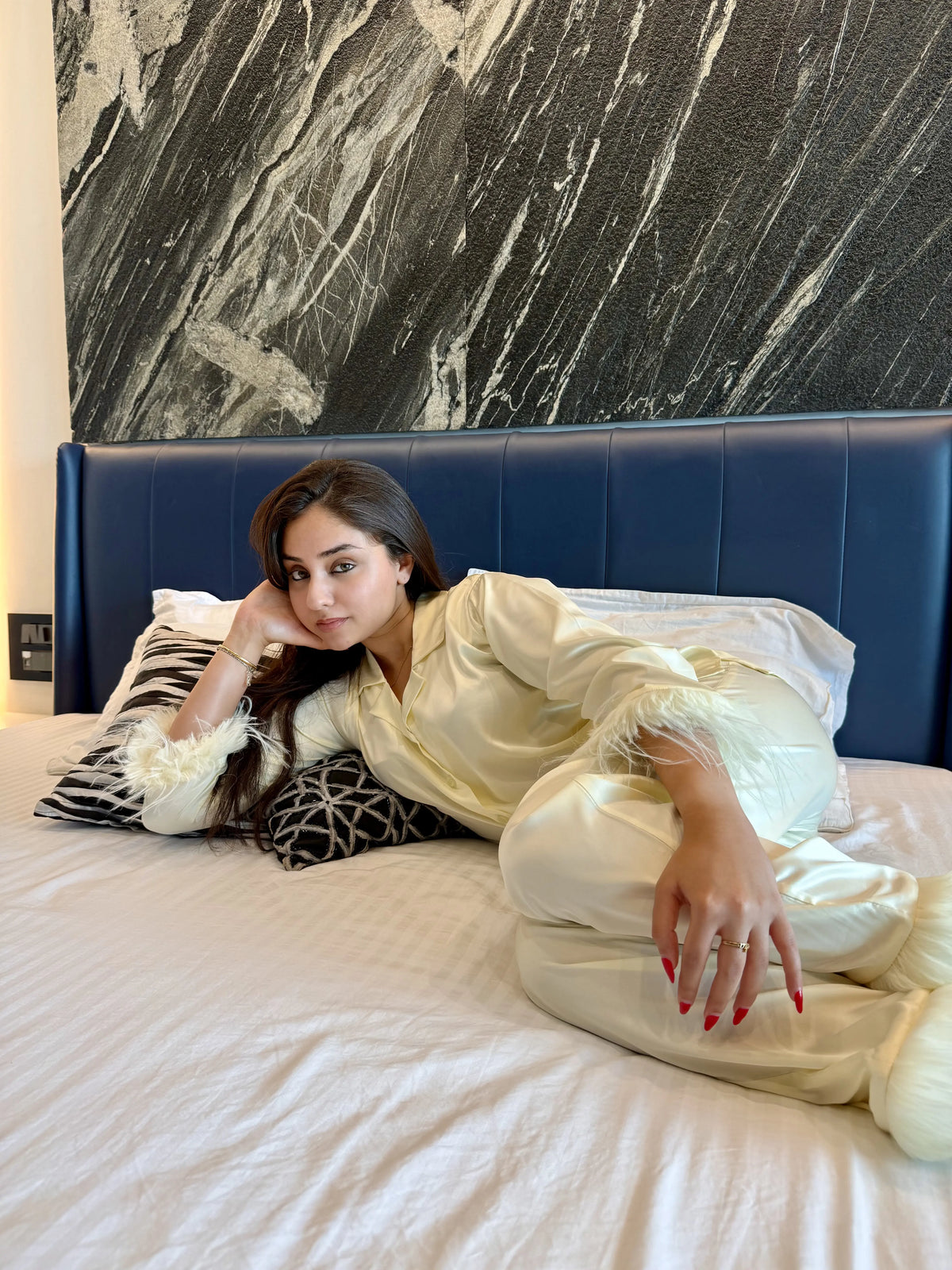 Woman in cream satin pajamas with feather details lying on white bed with blue headboard and black marble wall