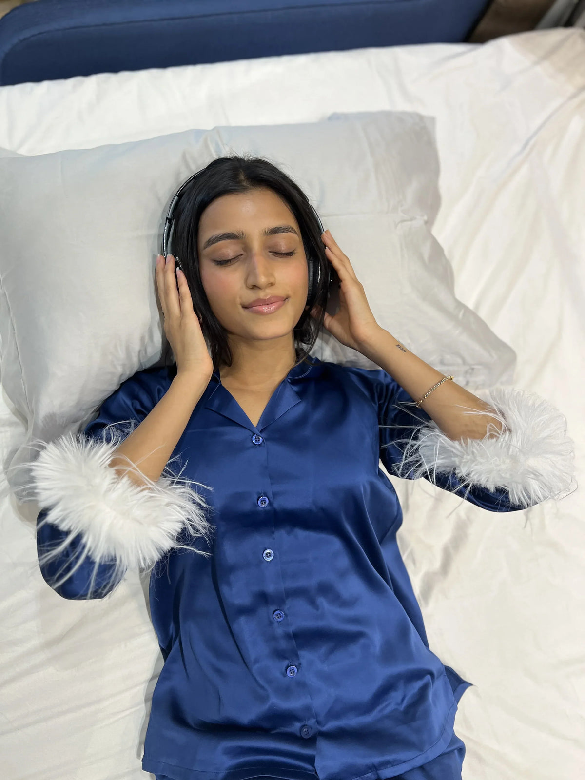 Relaxed woman in blue satin pajamas with white feather cuffs listening to music on headphones while lying on bed