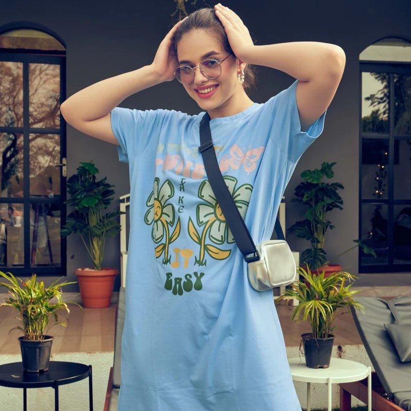 Smiling woman wearing oversized light blue floral printed dress with crossbody bag and glasses indoors