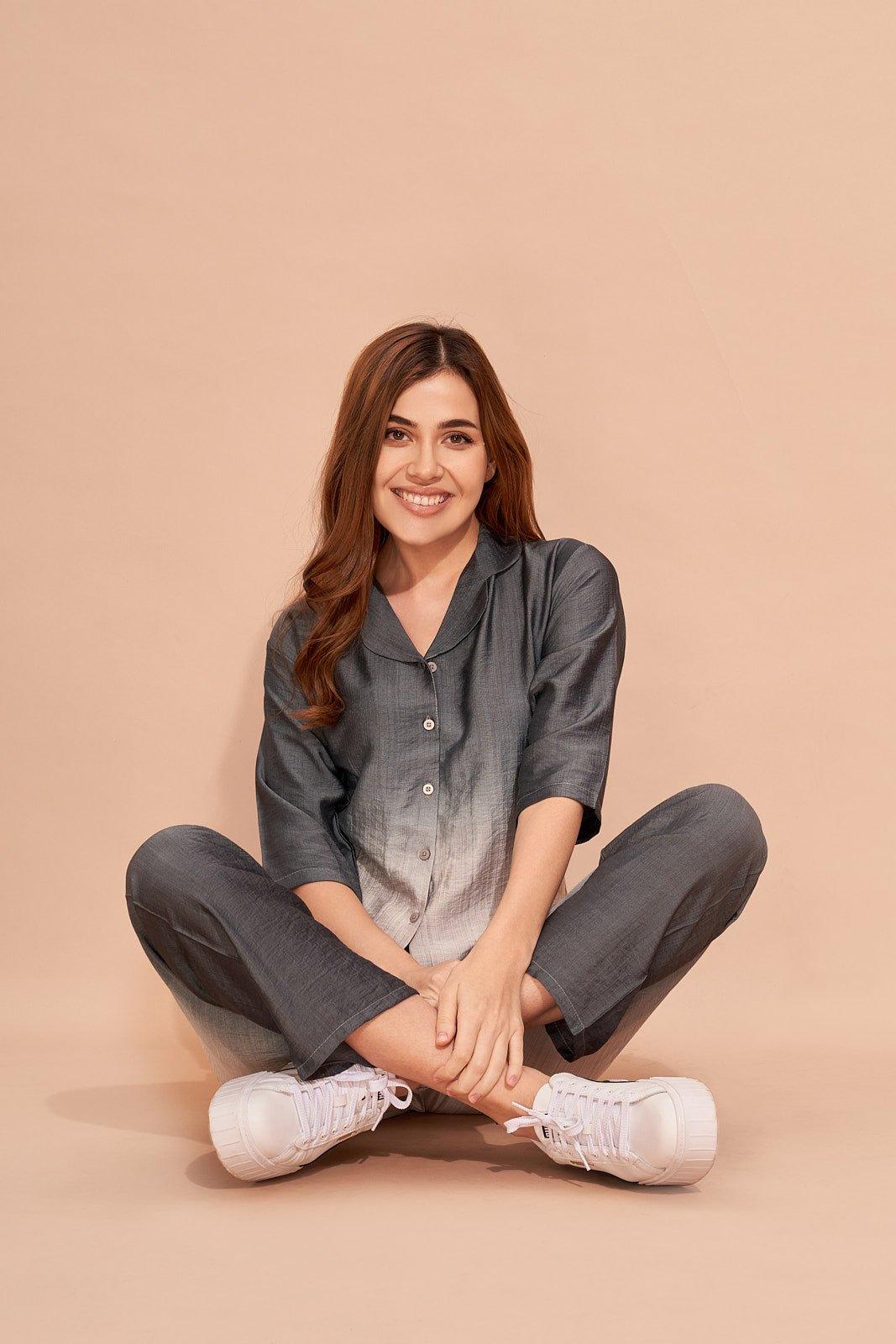 Smiling woman sitting cross-legged wearing a grey co-ord set with white sneakers on beige background