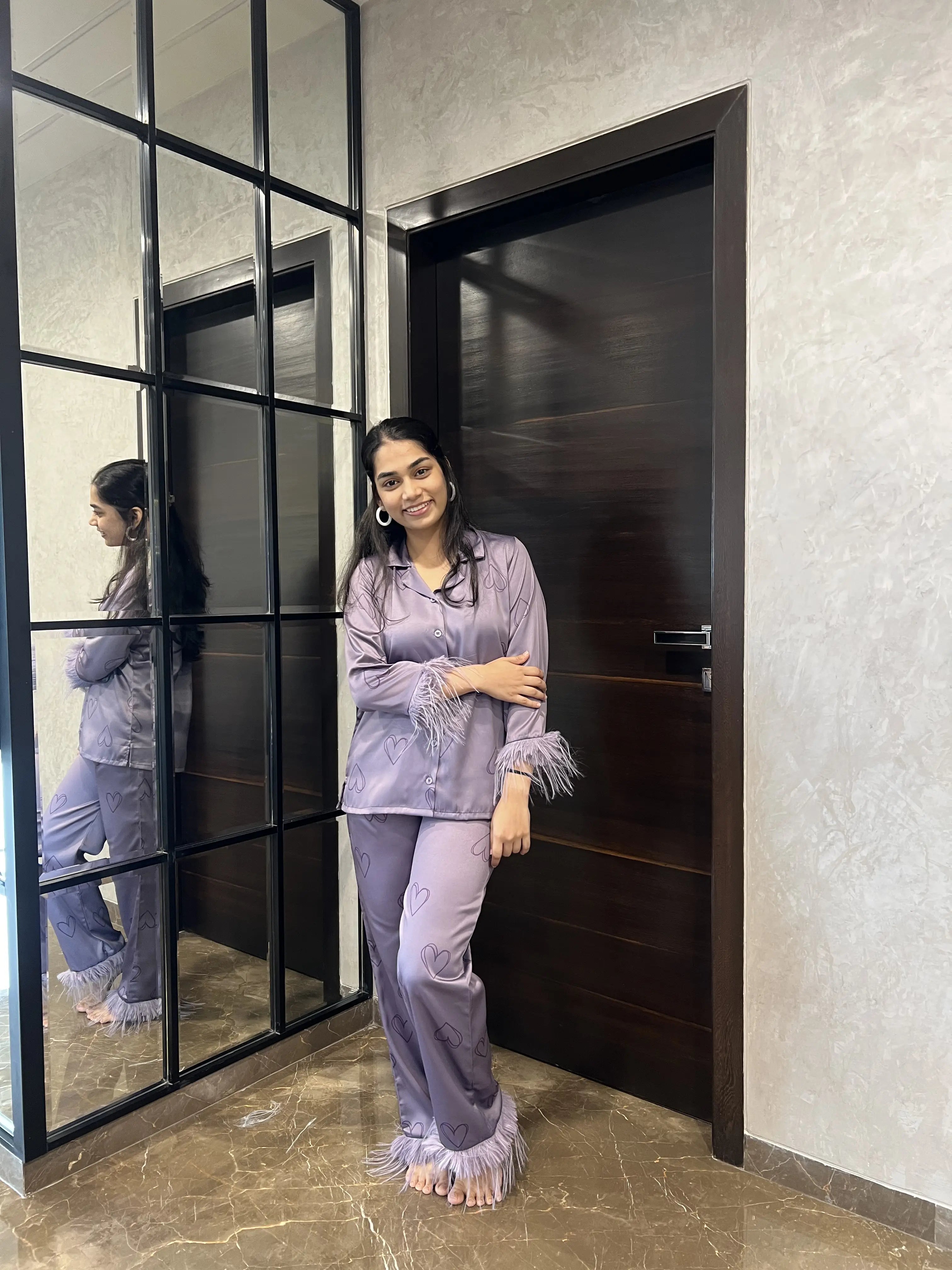 A woman in a lavender satin night suit is standing in front of a mirror in a room with a dark wooden door.