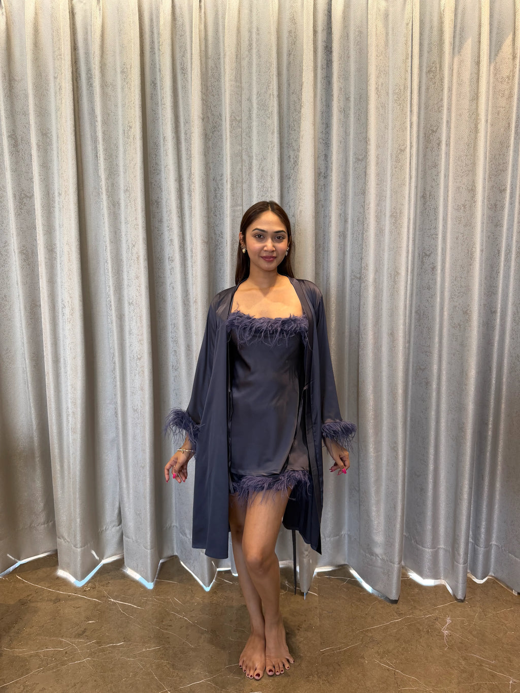 Woman wearing a dark grey satin nightgown with matching robe in front of a gray curtain.