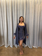 Woman wearing a dark grey satin nightgown with matching robe in front of a gray curtain.